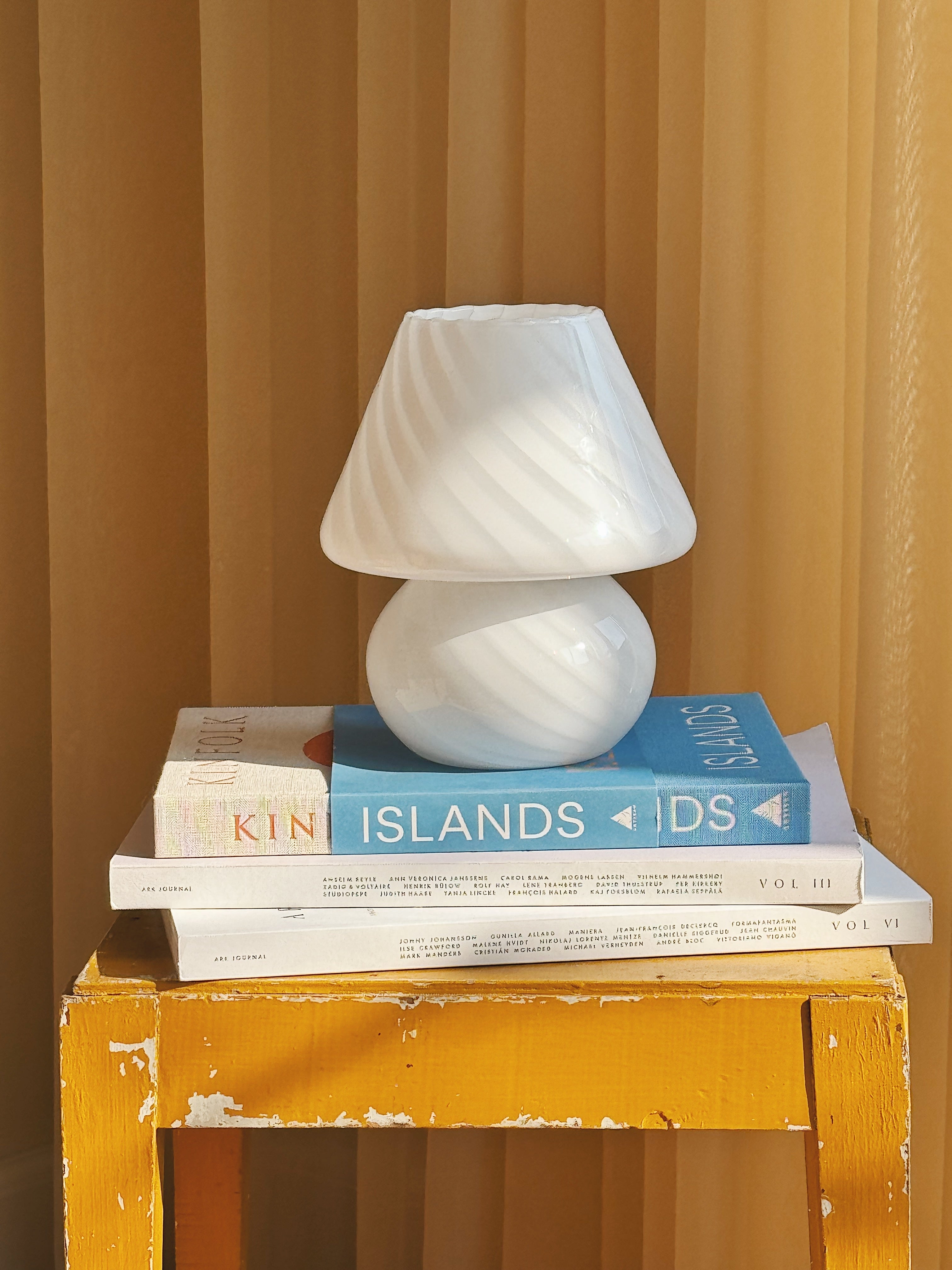 Vintage white Murano glass lamp with striped swirl design, styled on books atop a rustic yellow stool with warm curtain background