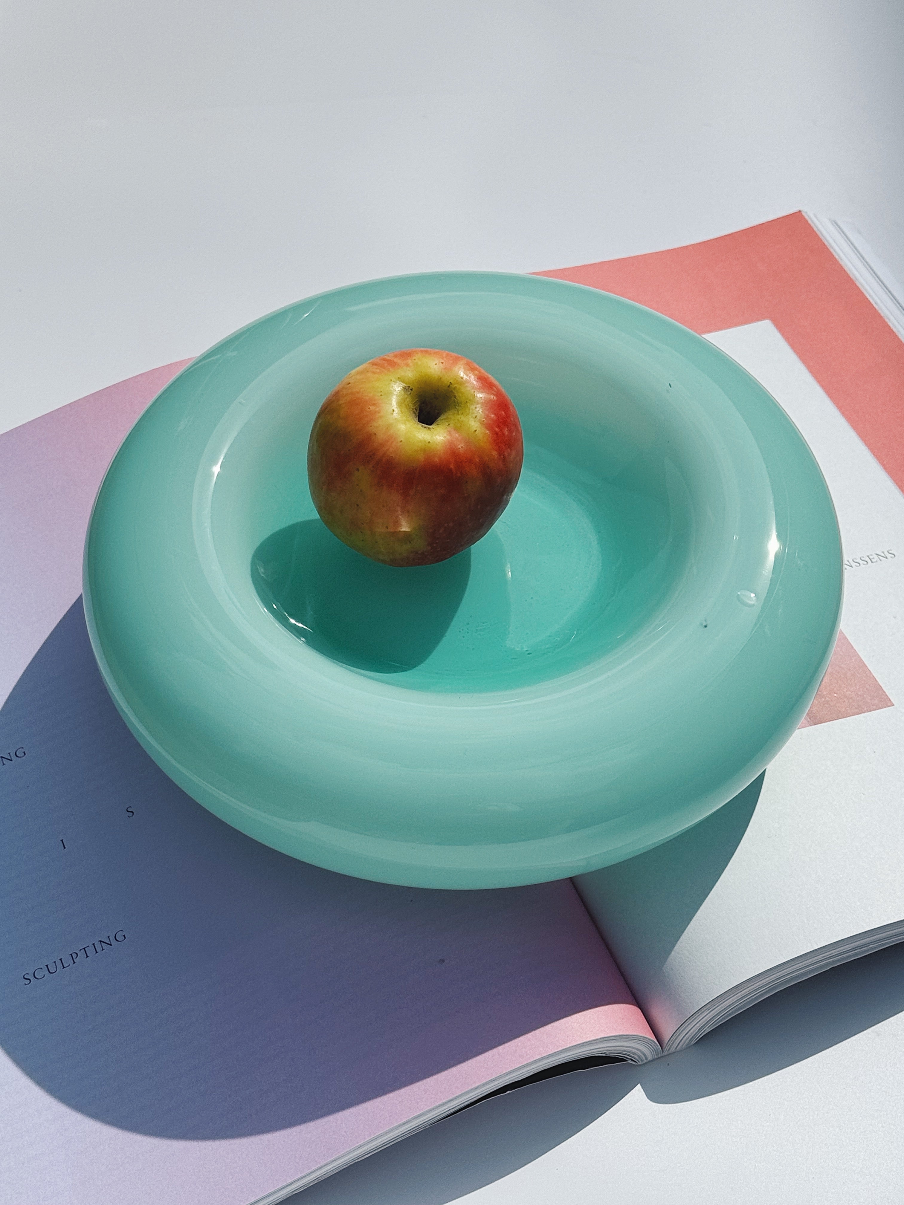 Turquoise Murano glass bowl styled with a red apple on an open magazine in sunlight – Studio Hafnia