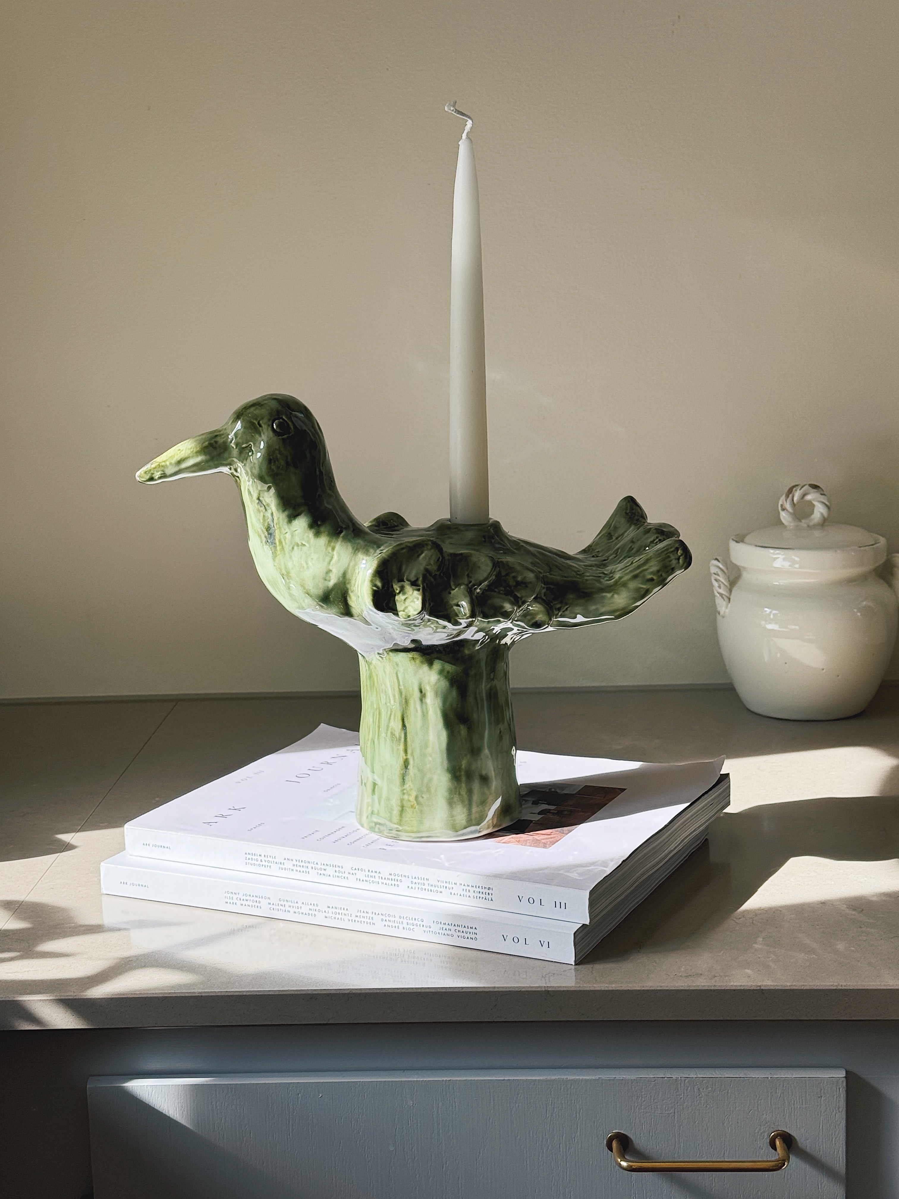 Green ceramic bird-shaped candlestick by Serax with a white taper candle, styled on books in natural sunlight