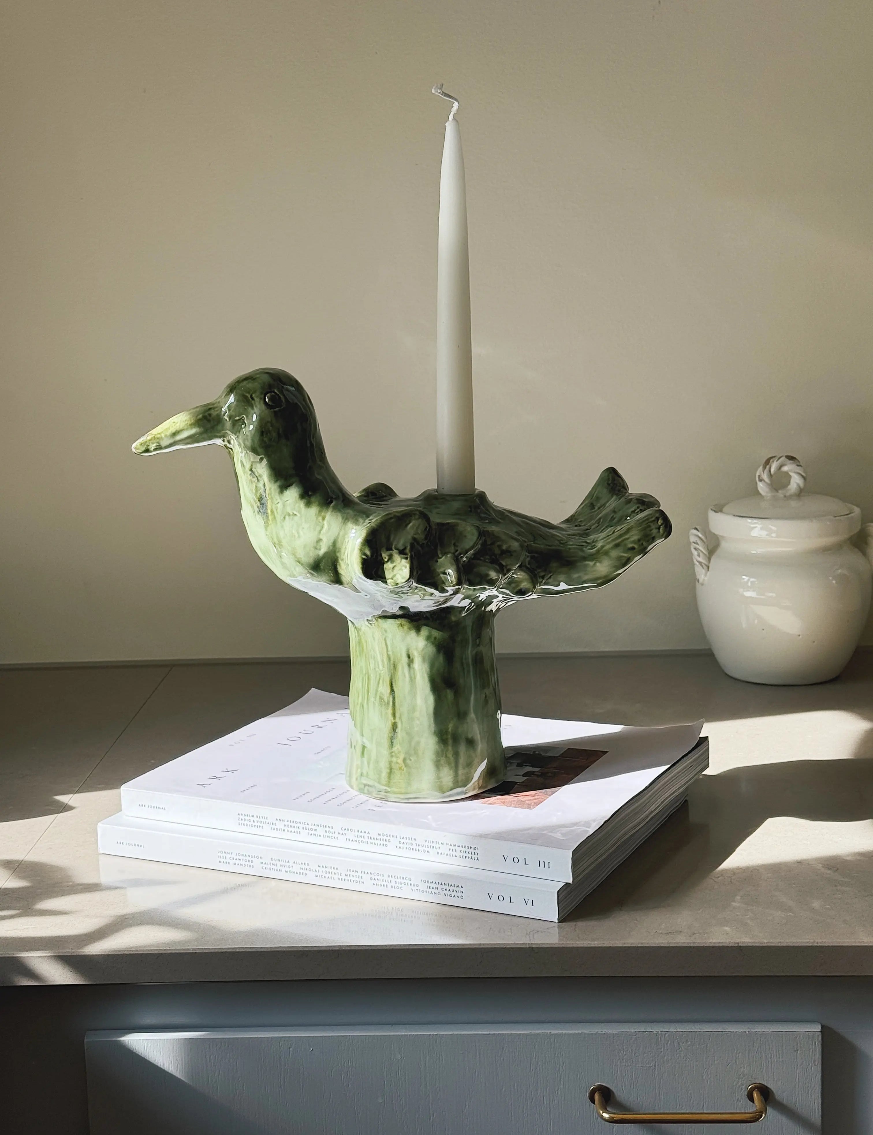 Green ceramic bird-shaped candlestick by Serax with a white taper candle, styled on books in natural sunlight