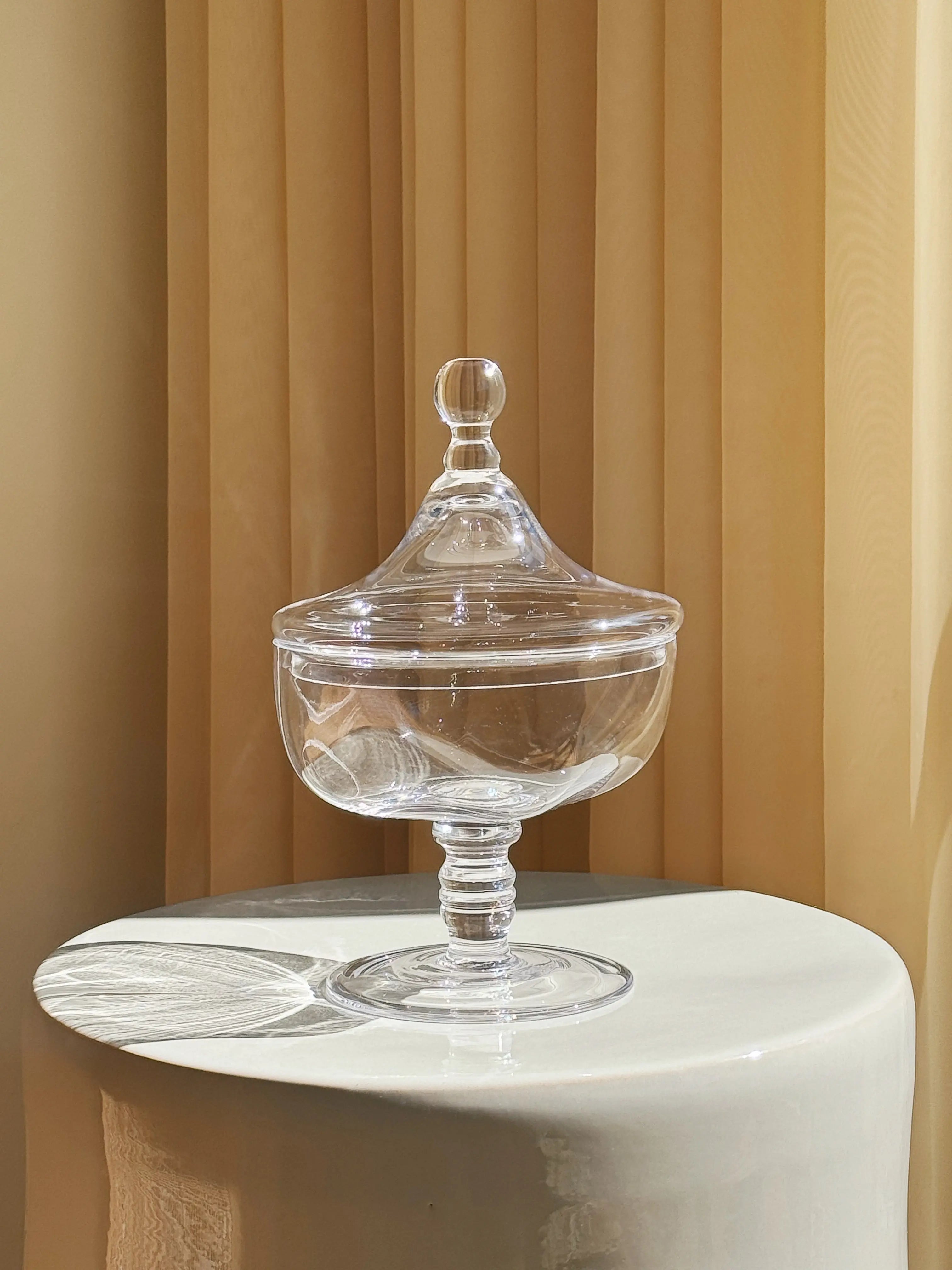 Josephine glass jar with lid by Serax, clear glass design displayed on pedestal with warm beige curtain background