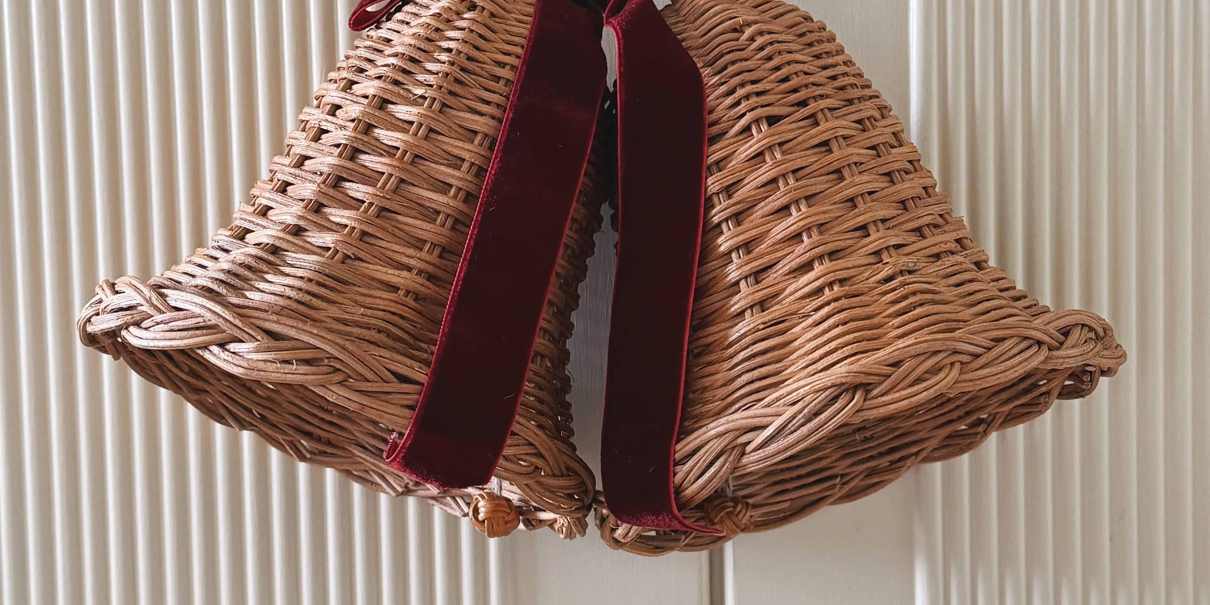 Pair of woven rattan Christmas bells tied with a deep red velvet ribbon, hanging on a white wooden door.