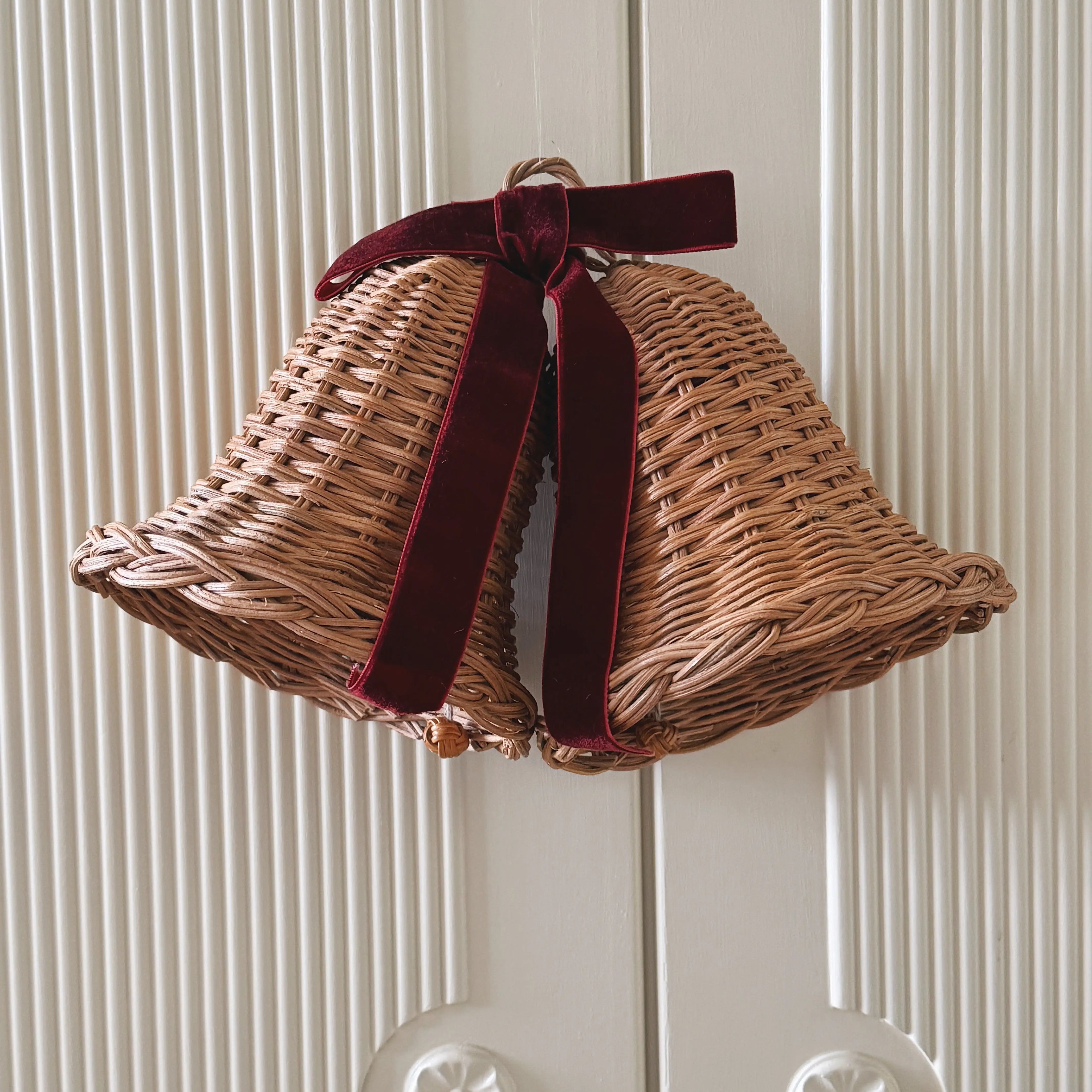 Pair of woven rattan Christmas bells tied with a deep red velvet ribbon, hanging on a white wooden door.