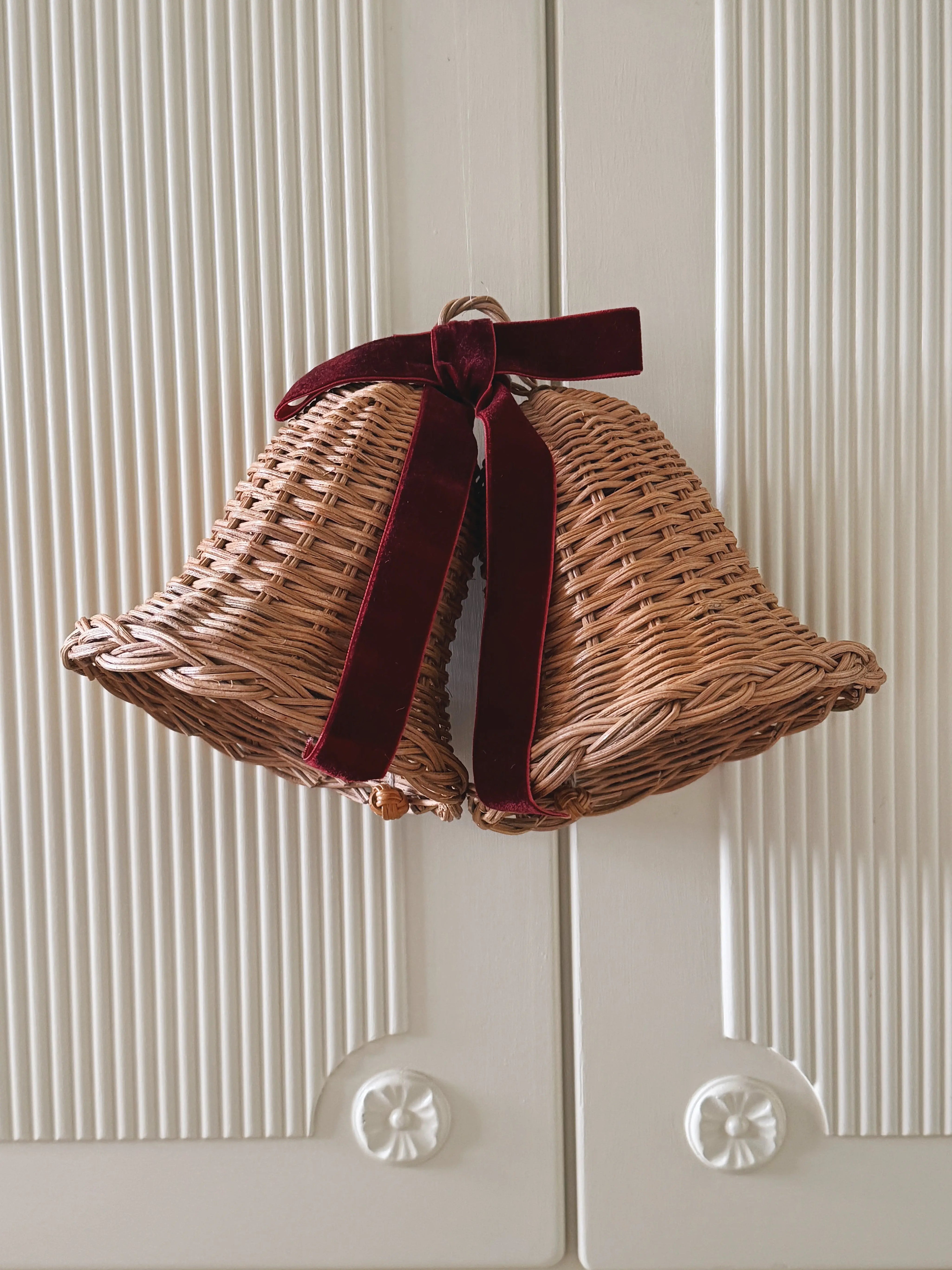 Pair of woven rattan Christmas bells tied with a deep red velvet ribbon, hanging on a white wooden door.