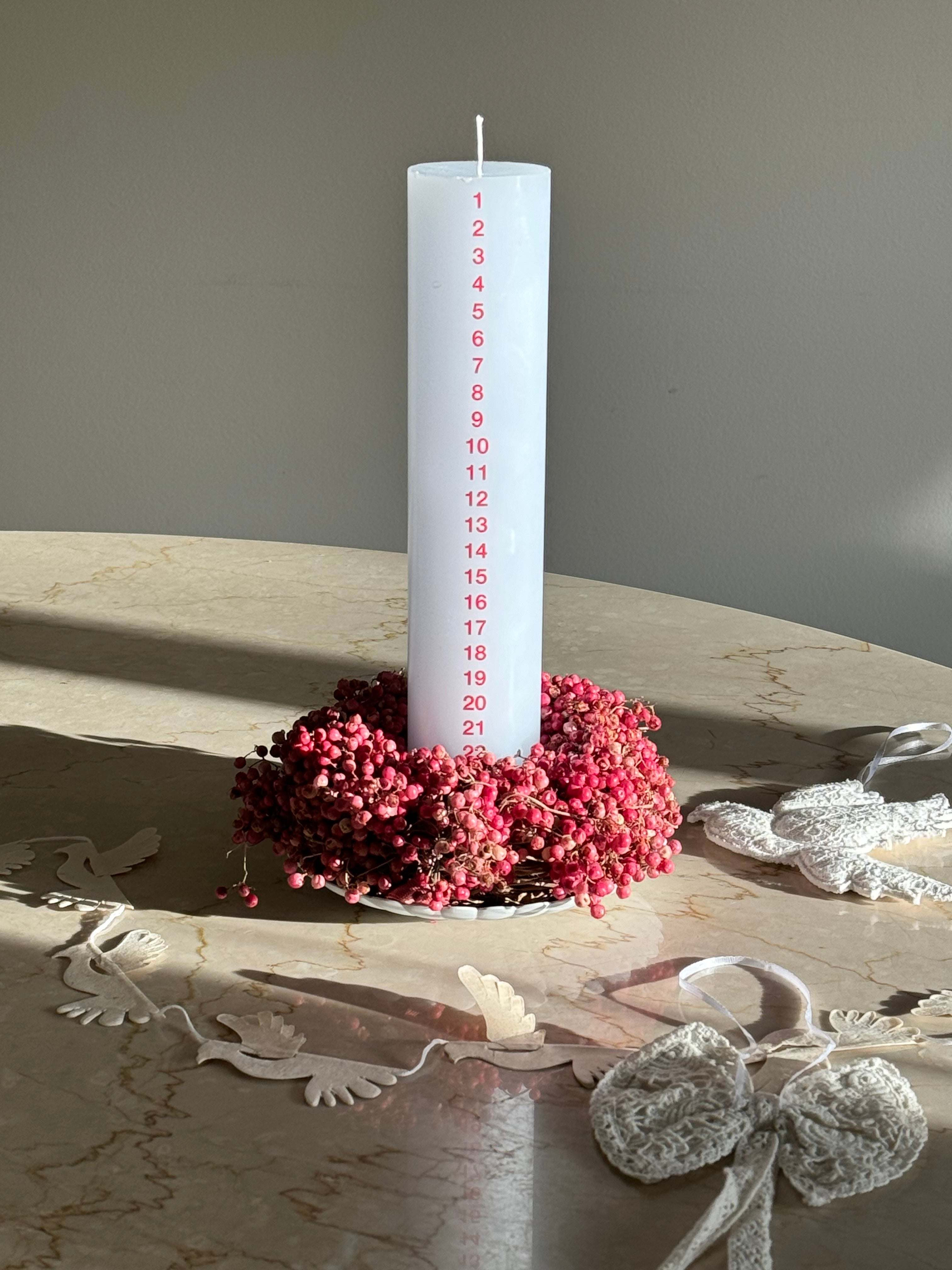 White advent candle in stearin from Kunstindustrien with red numbers, decorated with a wreath of red berries on marble table
