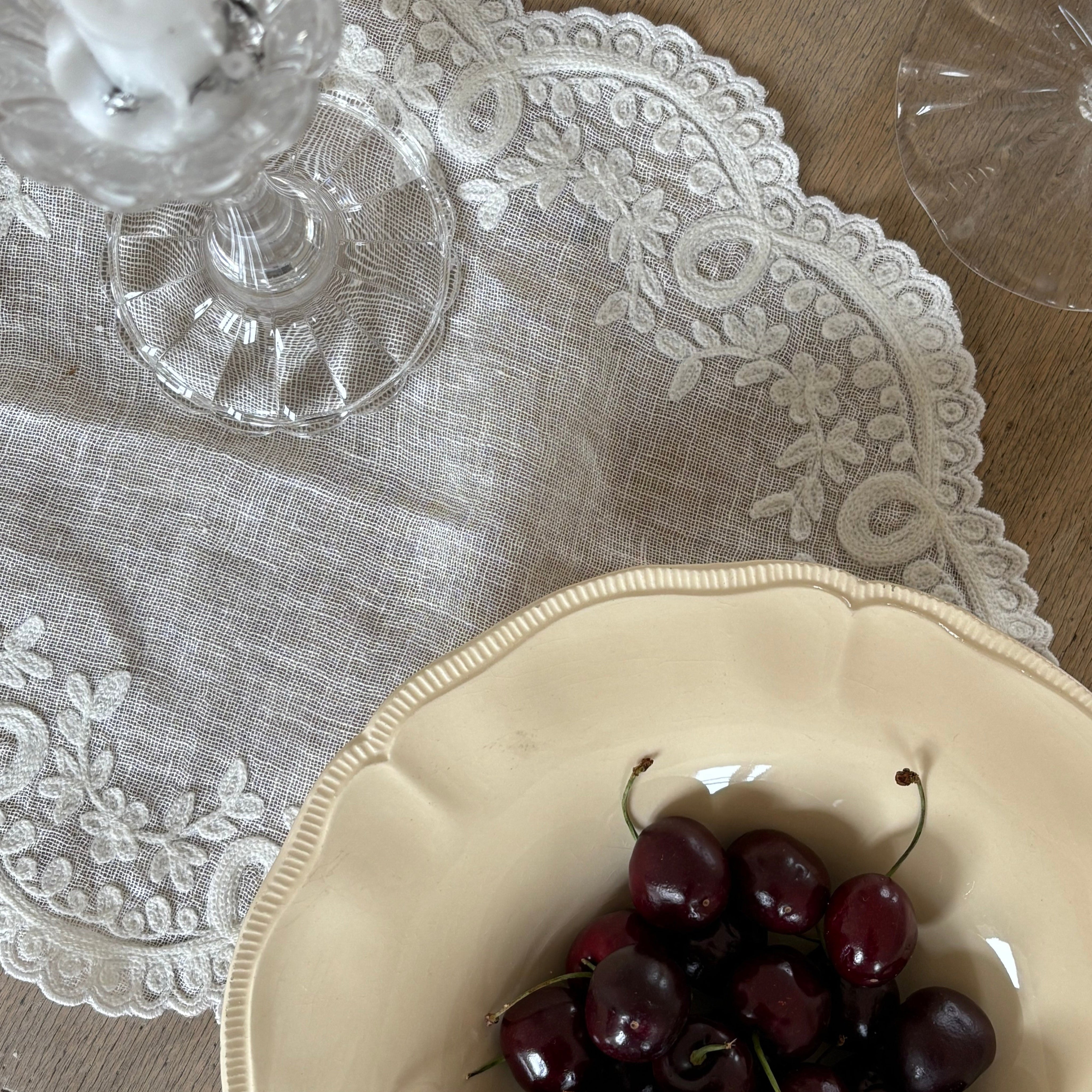 Elegant linen tablecloth with embroidered lace edge used as table textile, styled with glass candlestick, wine glass and ceramic bowl with cherries.