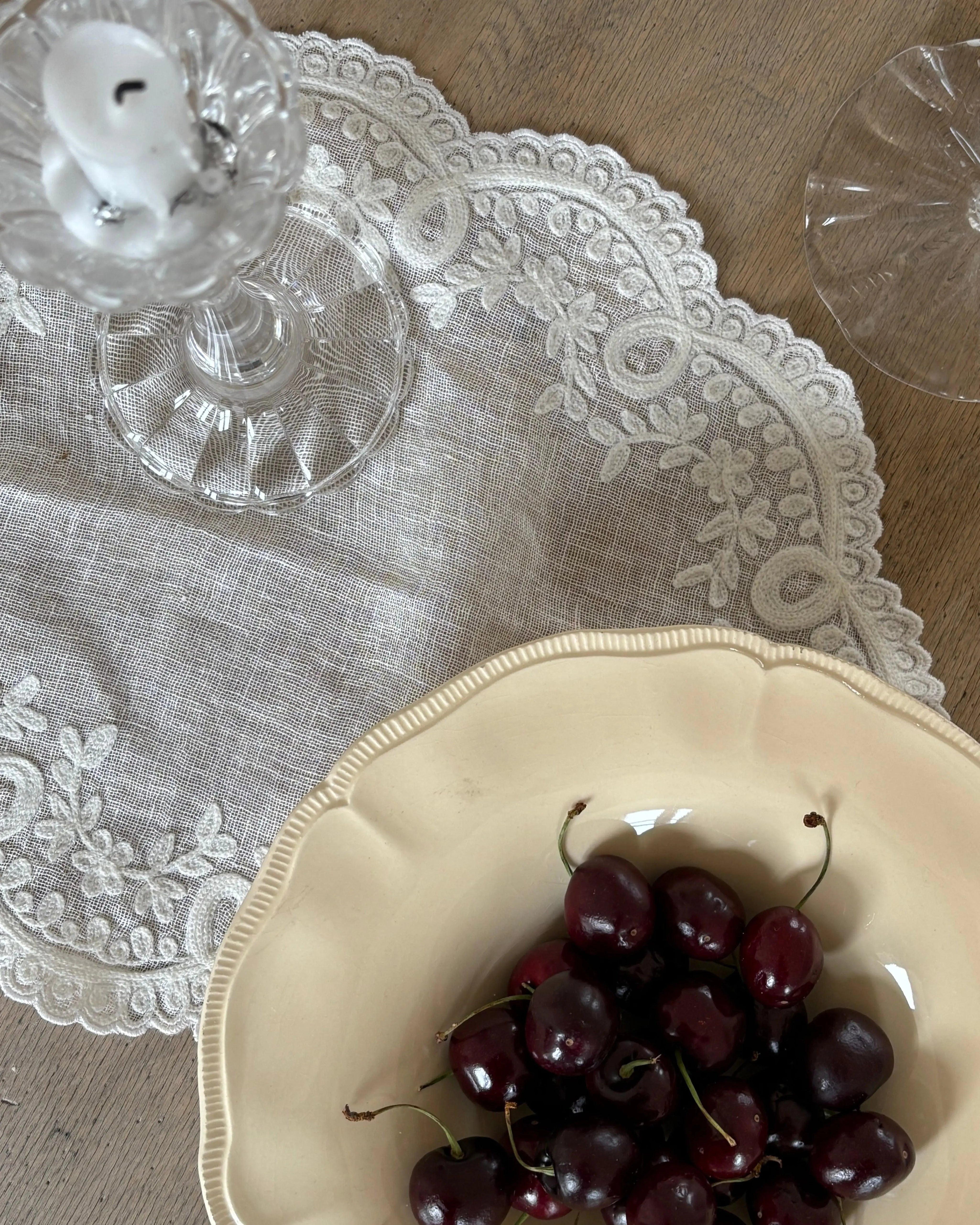Elegant linen tablecloth with embroidered lace edge used as table textile, styled with glass candlestick, wine glass and ceramic bowl with cherries.