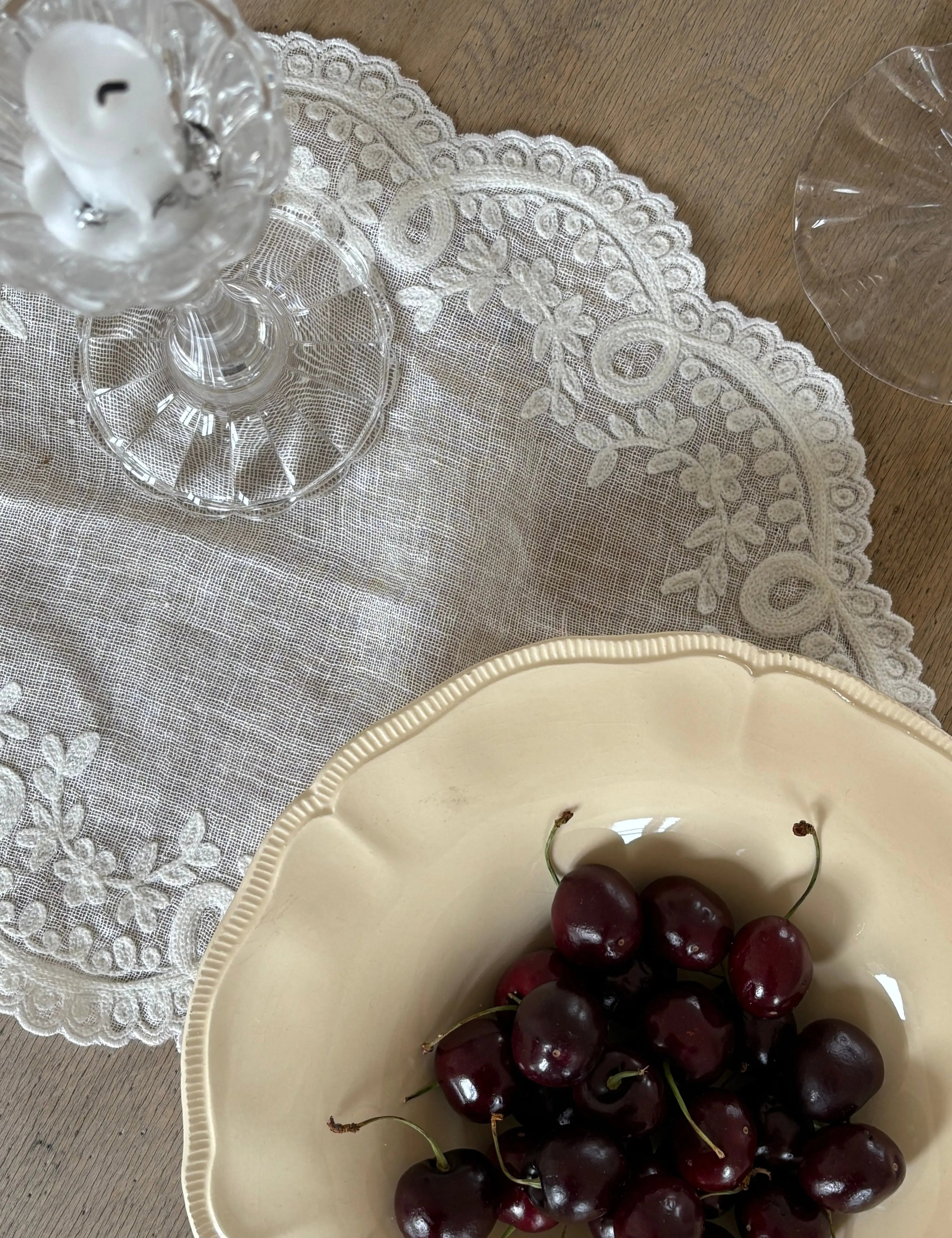 Elegant linen tablecloth with embroidered lace edge used as table textile, styled with glass candlestick, wine glass and ceramic bowl with cherries.