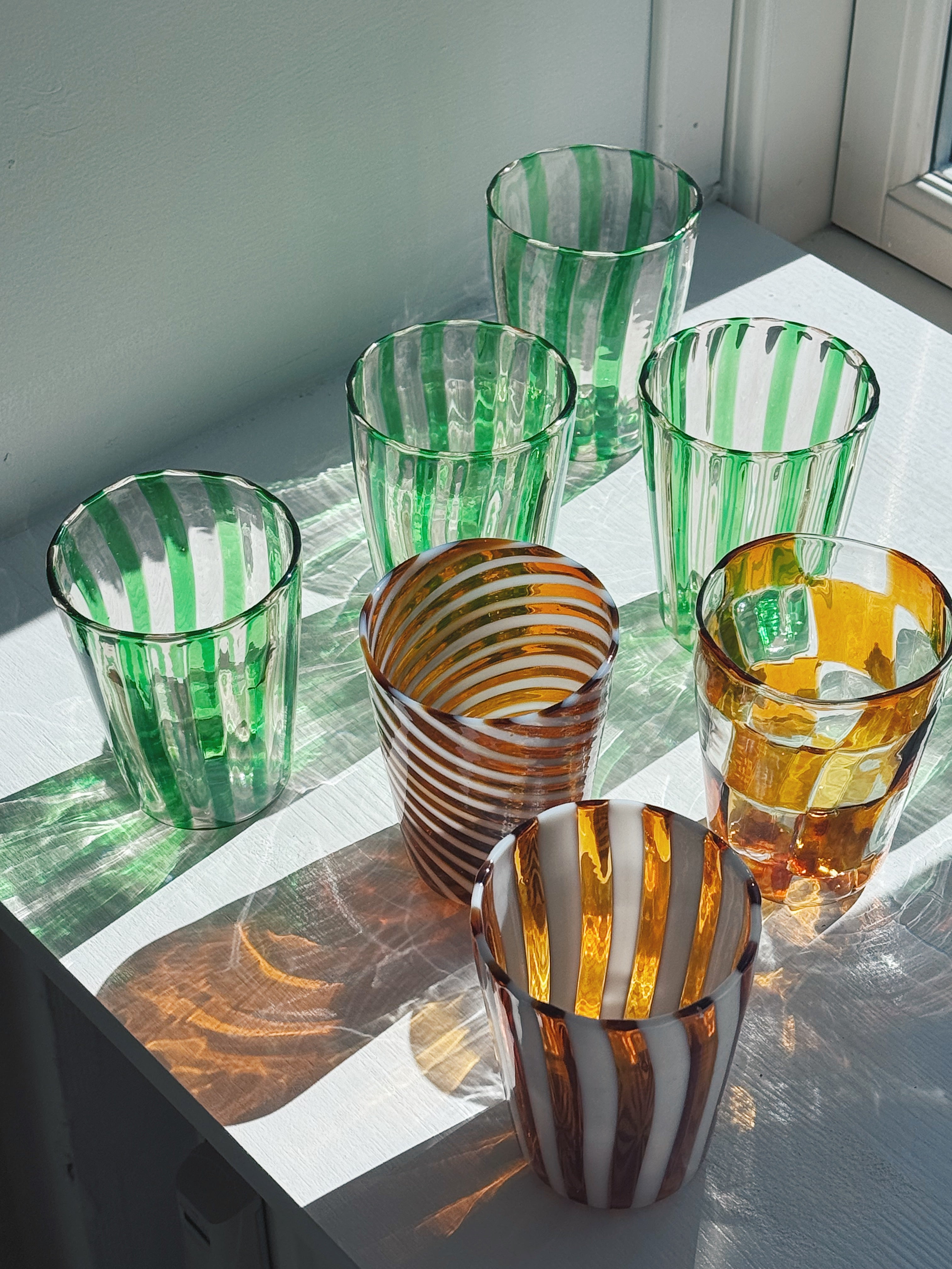 Håndblæste Murano drikkeglas i sollys på en vindueskarm – grønstribede glas, ravfarvet swirl og hvidstribet glas – Studio Hafnia