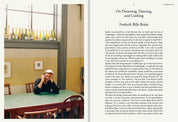 Frederik Bille Brahe at a green café table with wine bottles on the windowsill and the essay On Dreaming, Dancing, and Cooking