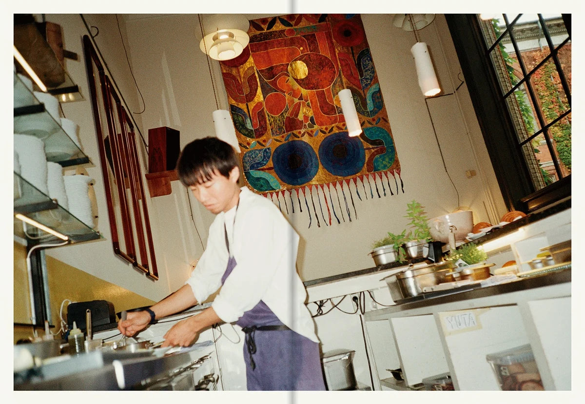 Chef in a white shirt and blue apron at the stove in the Apollo kitchen with a colourful woven wall hanging