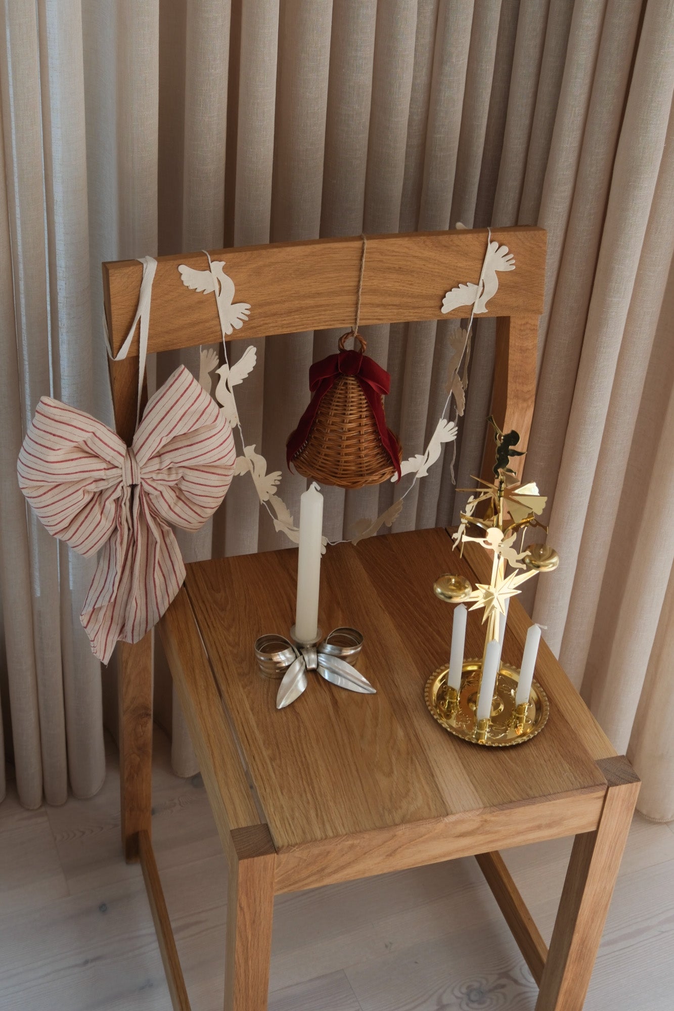 The five pieces of the Christmas package styled on an oak chair – hanging rattan bell, red and white striped bow, dove garland, silver candlestick and brass angel chime