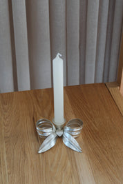 Candlestick shaped like a bow in matte silver with a white candle on an oak table