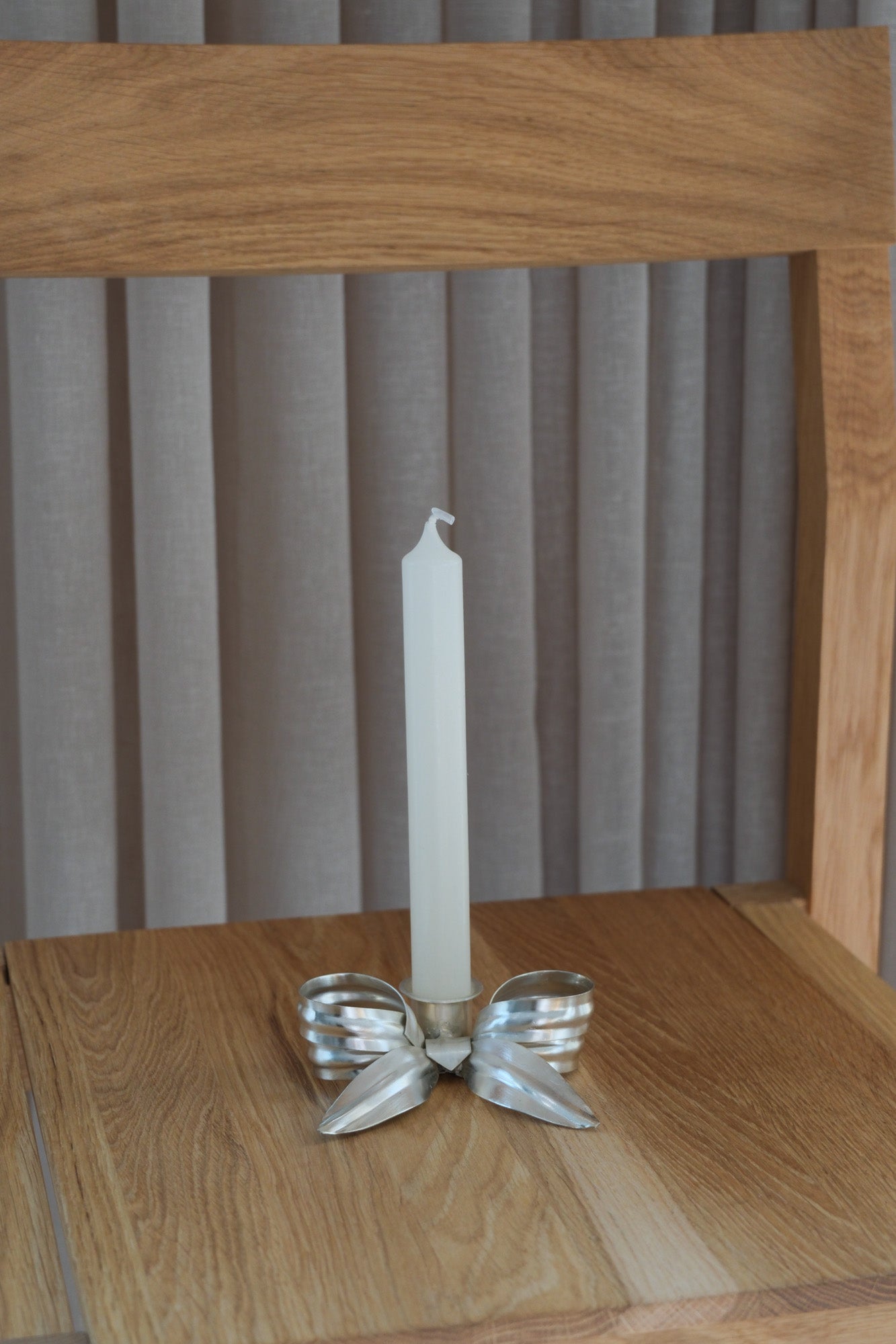 Bow-shaped candlestick in matte silver with a white candle, seen from the front on an oak chair