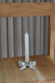 Bow-shaped candlestick in matte silver with a white candle, seen from the front on an oak chair