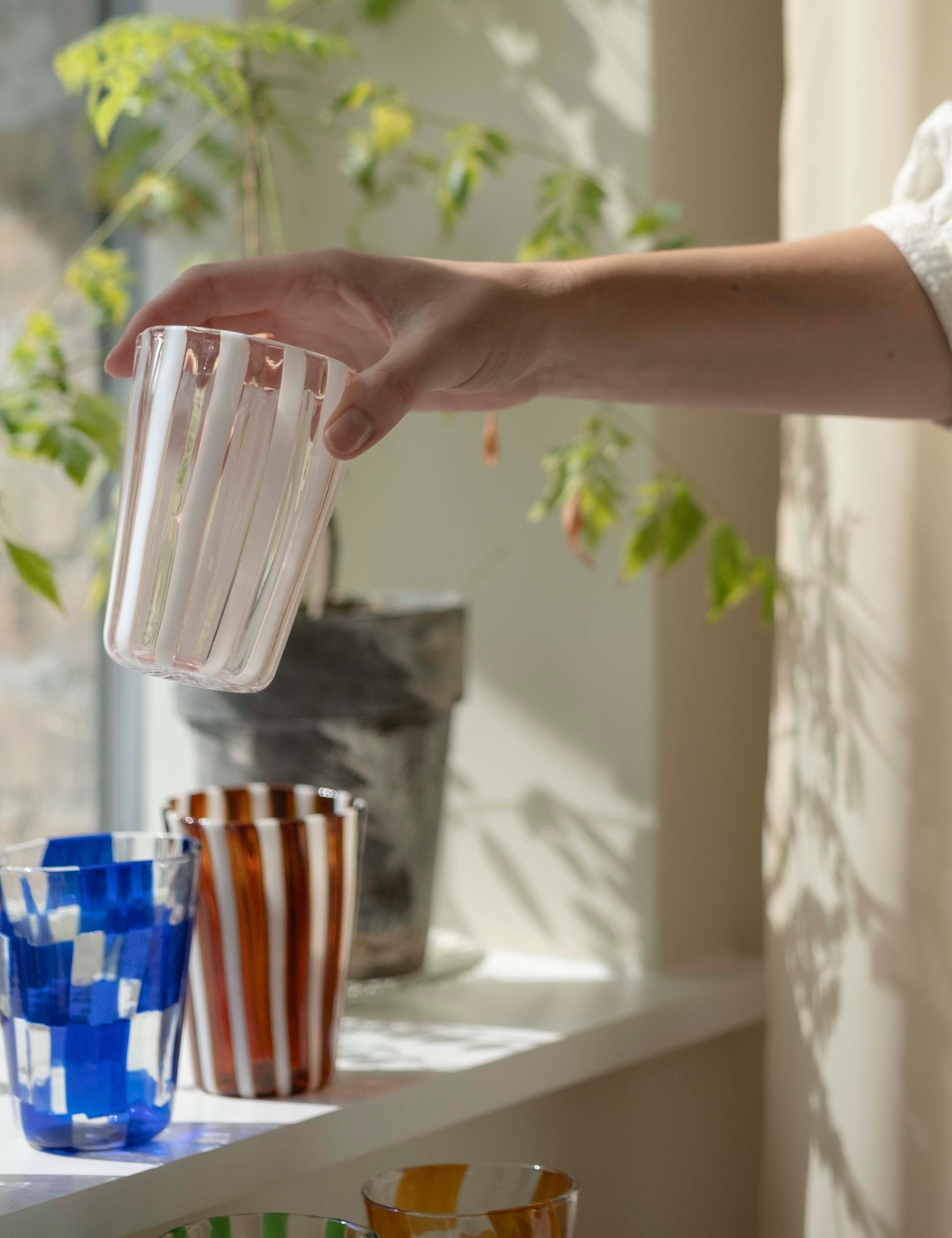 Hånd sætter et håndblæst Murano-glas med striber i vindueskarmen ved siden af farvede drikkeglas