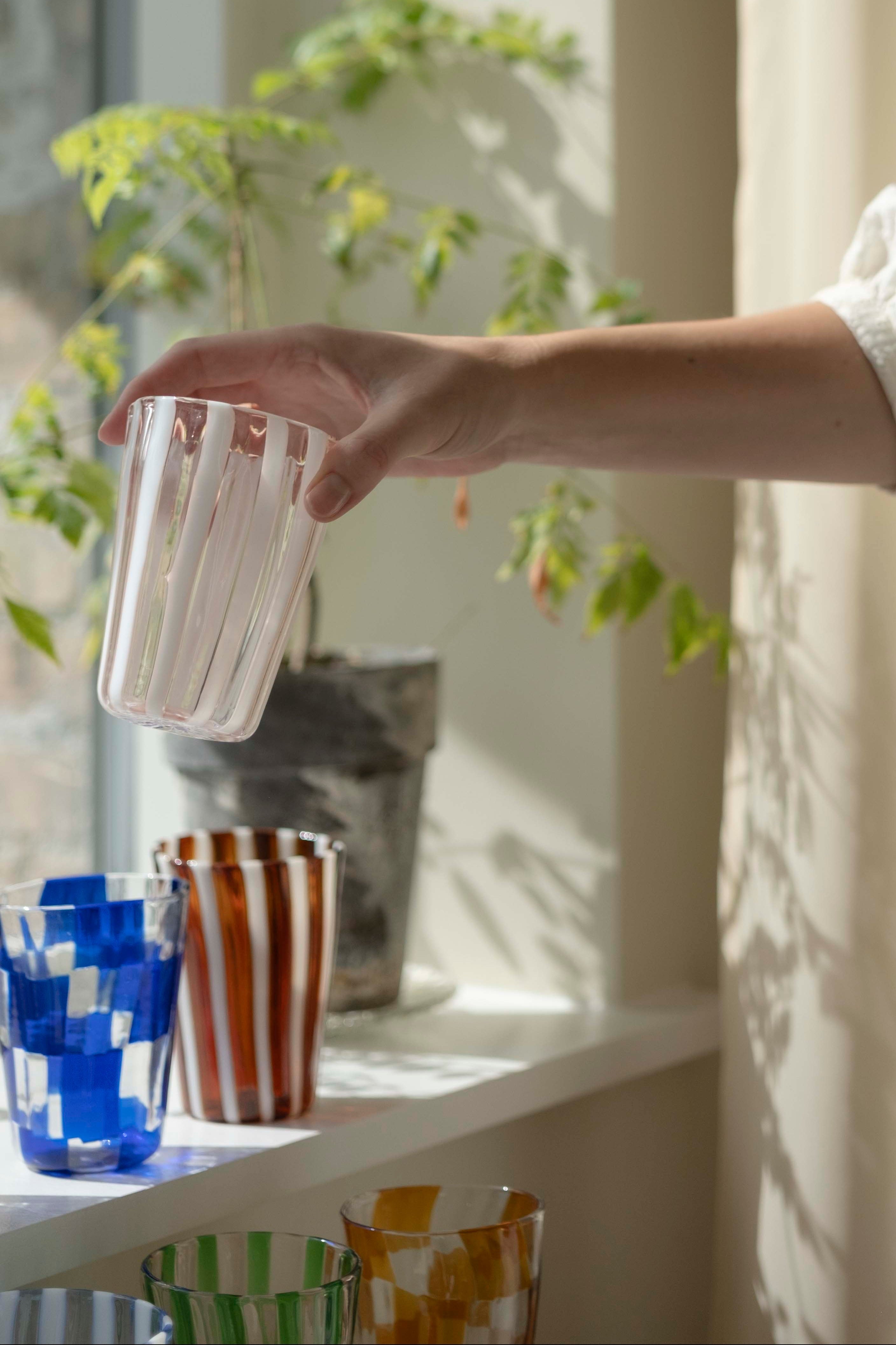 Hånd sætter et håndblæst Murano-glas med striber i vindueskarmen ved siden af farvede drikkeglas